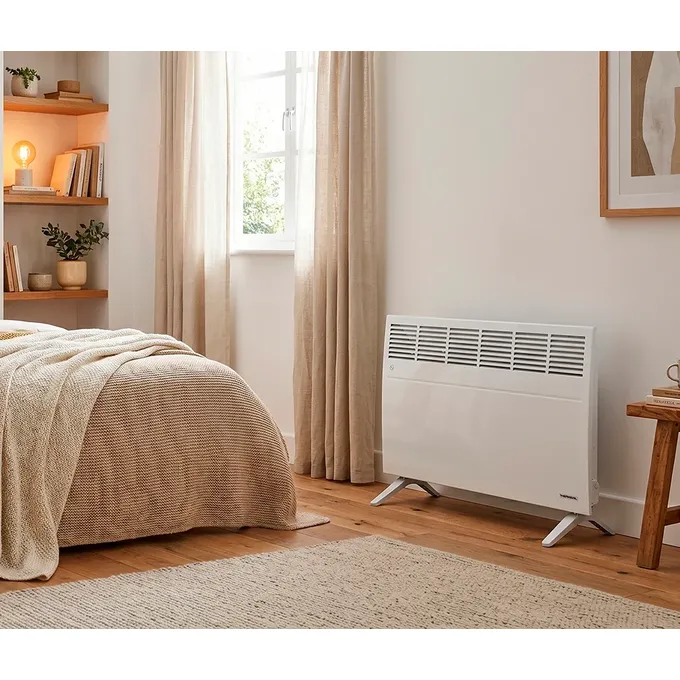 Biały grzejnik elektryczny Thermoval TX Next 2500W ustawiony na drewnianej podłodze w sypialni, obok łóżka.