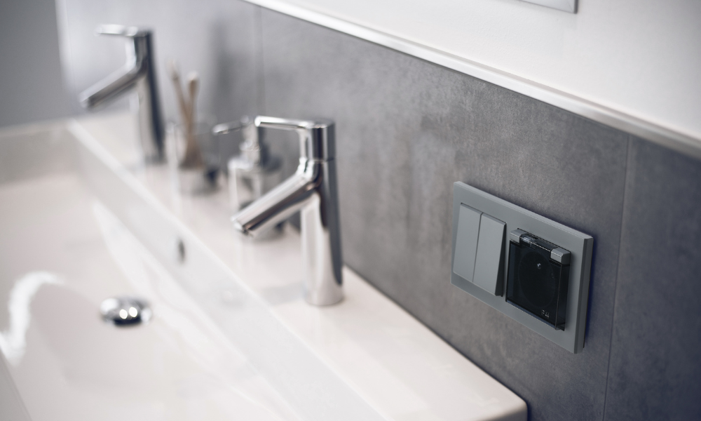 Łącznik i gniazdo do łazienki z klapką w kolorze Szary Grafit z serii Ospel Sonata na tle kafelków imitujących beton