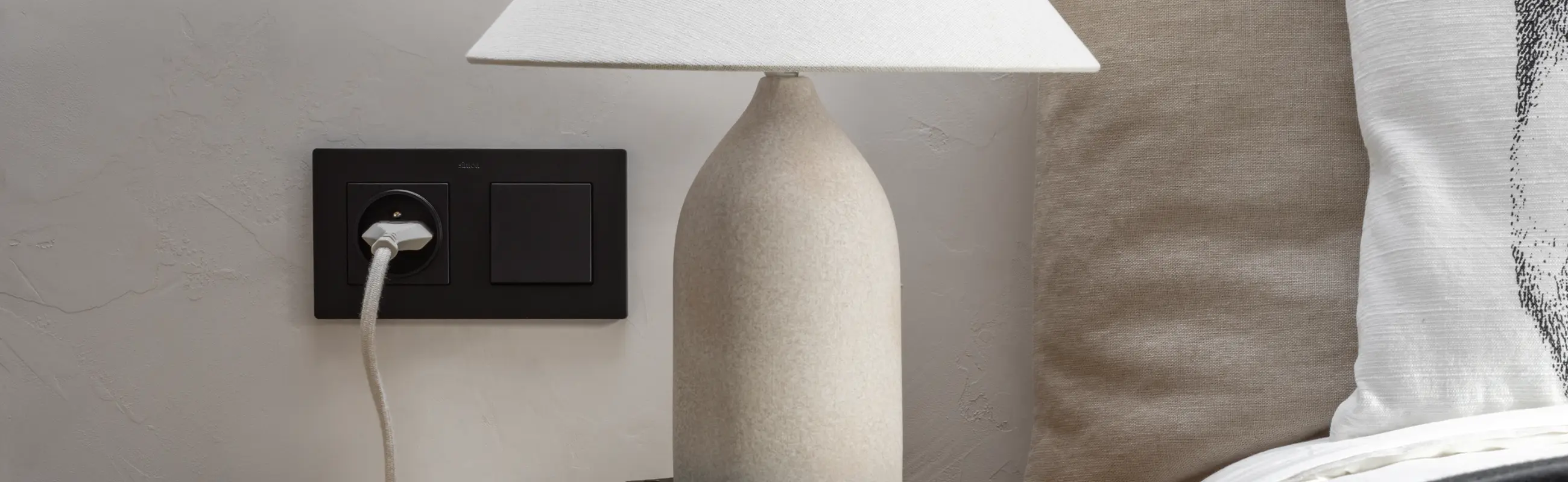 Czarne gniazdo z wpiętą wtyczką i czarny włącznik w podwójnej ramce przy ceramicznej lampce nocnej